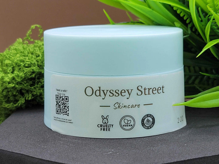 Skincare jar with 'Odyssey Street' branding on a dark surface with green foliage in the background